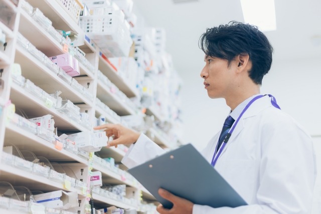 ワイシャツ・ネクタイ・白衣を着用している薬剤師男性が左手にバインダーを持ち、右手で調剤室にある調剤棚から包装されている医薬品を取り出そうとしている画像