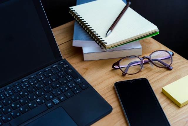 ノートパソコンの横に、スマートフォン、眼鏡、メモ帳、ノートとペンが並べられた木製デスクの上の作業スペース