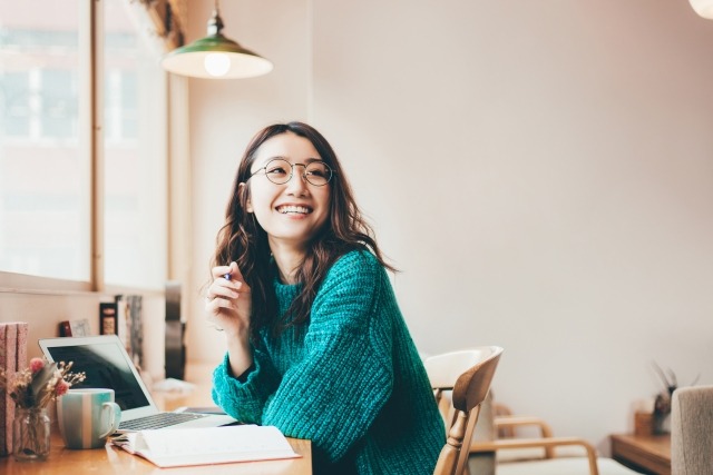 カフェのような明るい空間で、眼鏡をかけた女性がノートとノートパソコンの前に座り、振り返りながら笑顔を見せている様子