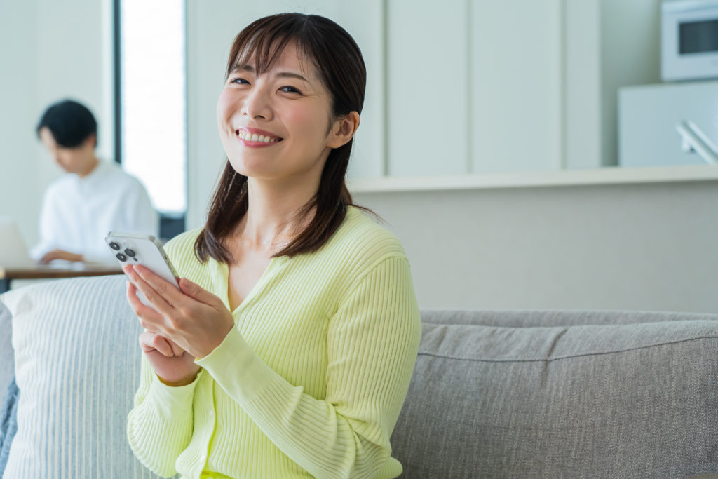 自宅のソファで笑顔を浮かべながらスマホを操作する女性