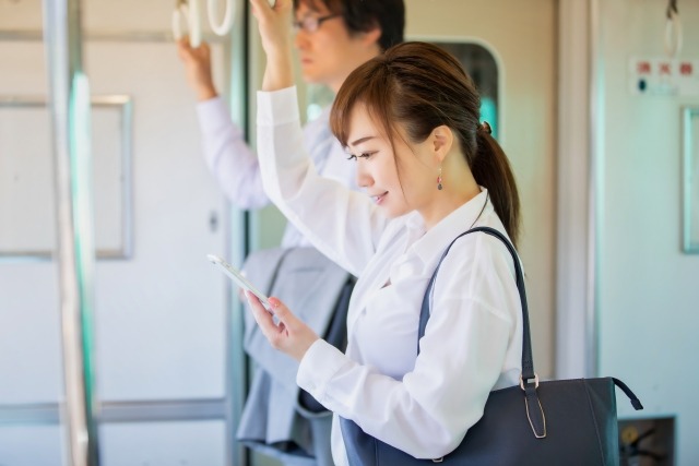 女性が電車の中で、吊革につかまり立ちしながらスマホを見て微笑んでいる