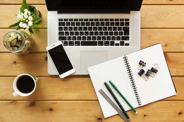 テーブルに、ノートパソコン、ノート、スマホ、コーヒーなどが置かれている