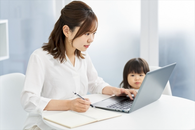 女性が真剣な顔でパソコン作業をしている。近くで小さな子どもが見ている。